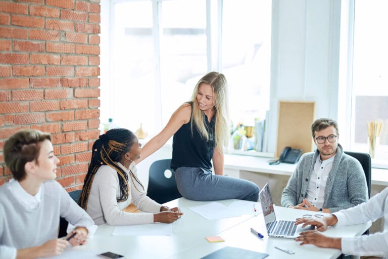 Vier Personen diskutieren an einem Tisch in einem modernen Büro. Die Hand der Chefin ruht auf der Schulter einer ihrer Mitarbeiterinnen, während sie mit ihr ermutigend spricht. Die Chefin symbolisiert ein fürsorgendes Eltern-Ich aus der Transaktionsanalyse.