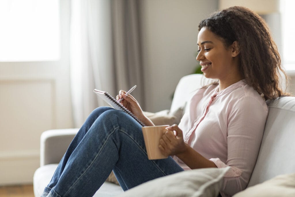 Eine Frau sitzt mit einer Kaffeetasse auf einem Sofa und schreibt ihren Lebensplan in ein Notizbuch.