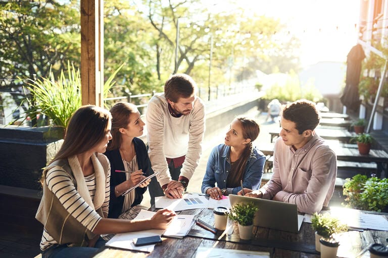 Eine Gruppe von Kollegen hält eine Besprechung draußen in einem Café. Symbolisch für VOPA+ Modell: Vernetzung, Offenheit, Partizipation, Agilität plus Vertrauen.