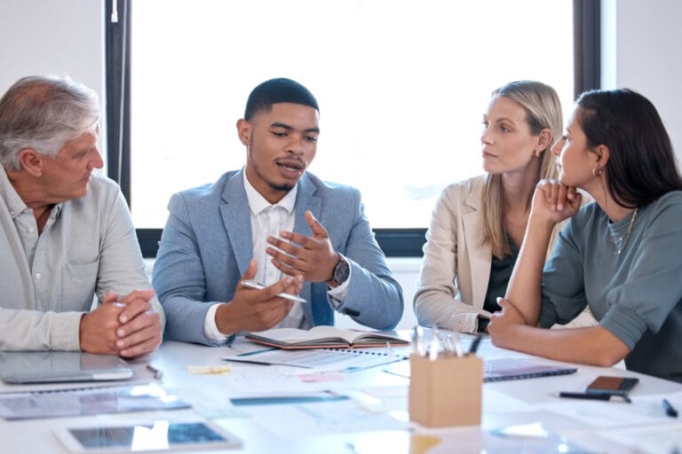 Ein HR-Coach sitzt mit vier Klienten an einem Besprechungstisch und stellt Fragen. Sinnbildlich: Coaching Ausbildung für HR-Coaches.